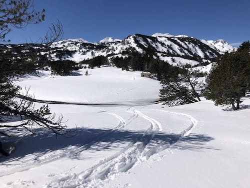 Descente à ski de randonnée dans la vallée du Galbe près de Font-Romeu, au départ de Formiguères, lors d'un raid, avec des traces de skis dans la neige, au milieu d'un paysage de sapins enneigés et de sommets, et une cabane en pierre visible au loin.