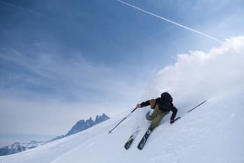 Skieur en pleine session de freeride, effectuant un virage dynamique et projetant de la neige derrière ses spatules, un beau ciel bleu et des montagnes au loin.