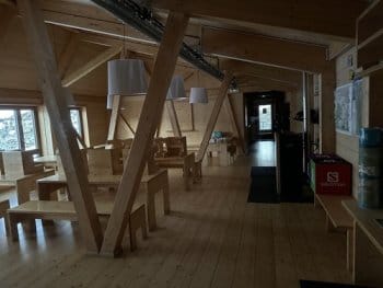 Intérieur en bois du refuge de l'Illa en Andorre, avec des tables en bois dans la salle à manger spacieuse à l'entrée. Un poêle réchauffe l'espace, tandis que des baies vitrées offrent une vue imprenable sur les sommets environnants Intérieur en bois du refuge de l'Illa en Andorre, avec des tables en bois dans la salle à manger spacieuse à l'entrée. Un poêle réchauffe l'espace, tandis que des baies vitrées offrent une vue imprenable sur les sommets environnants